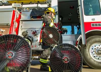 A firefighter in full protective gear shown from a low angle, about to step into a fire truck, holding BlowHard's Squirt PPV fan model over their right shoulder and looking away from the camera. The Quickee and Commando Models sit in front of him on the ground.