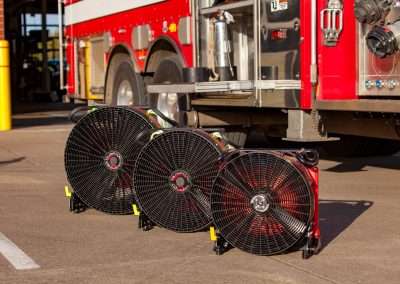 BlowHard's Commando, Quickee, and Squirt models lined up in that order, sit on the ground in front of a firetruck at a fire station. The fans are lined up from largest to smallest, showcasing their size comparison.
