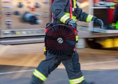 A firefighter in front of a fire truck carrying BlowHard's Squirt fan model over their right shoulder.