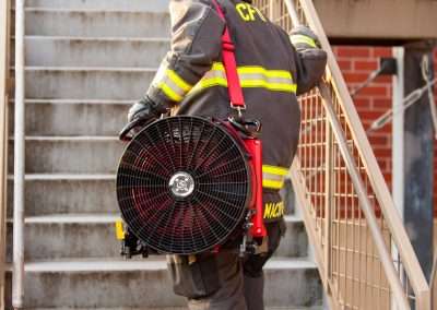 A firefighter walking up a staircase with the Squirt BlowHard fan model strapped over his left shoulder.