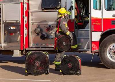 A firefighter in full protective gear about to step into a fire truck, holding BlowHard's Squirt PPV fan model over their right shoulder and looking in towards the firetruck. The Quickee and Commando Models sit in front of him on the ground.